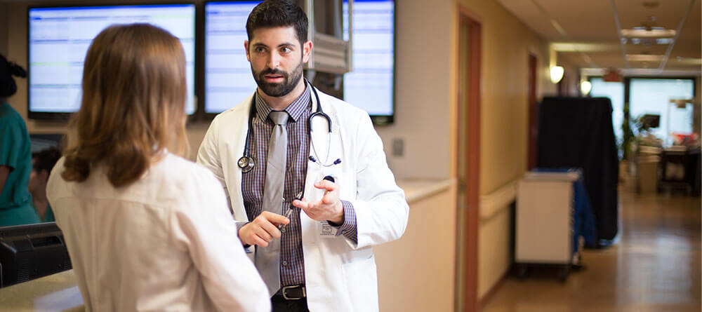 Male hospitalist speaks with another health care provider in hallway.